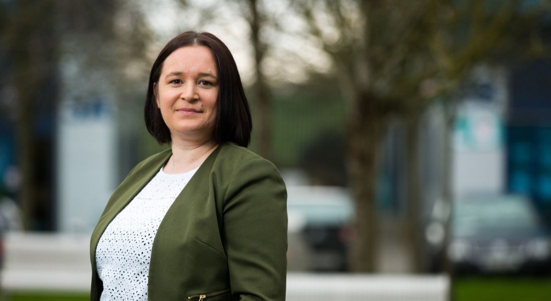 Dark-haired woman in green coat with white blouse standing in front of trees.