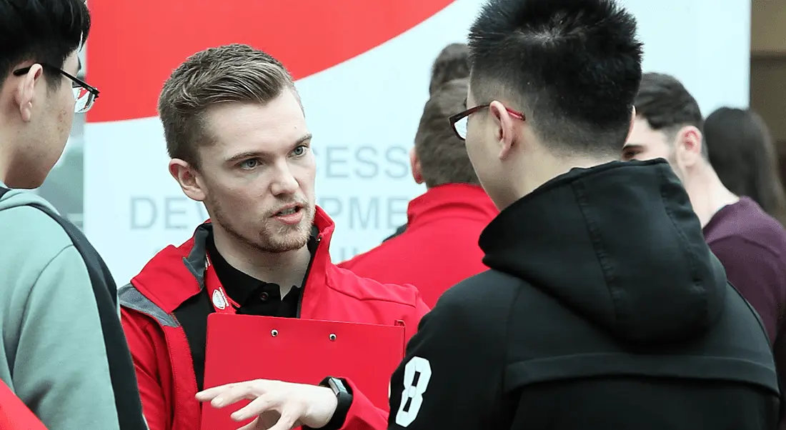 A man in a red windbreaker emblazoned with Takeda talking to fellow scientists in an open-plan building.