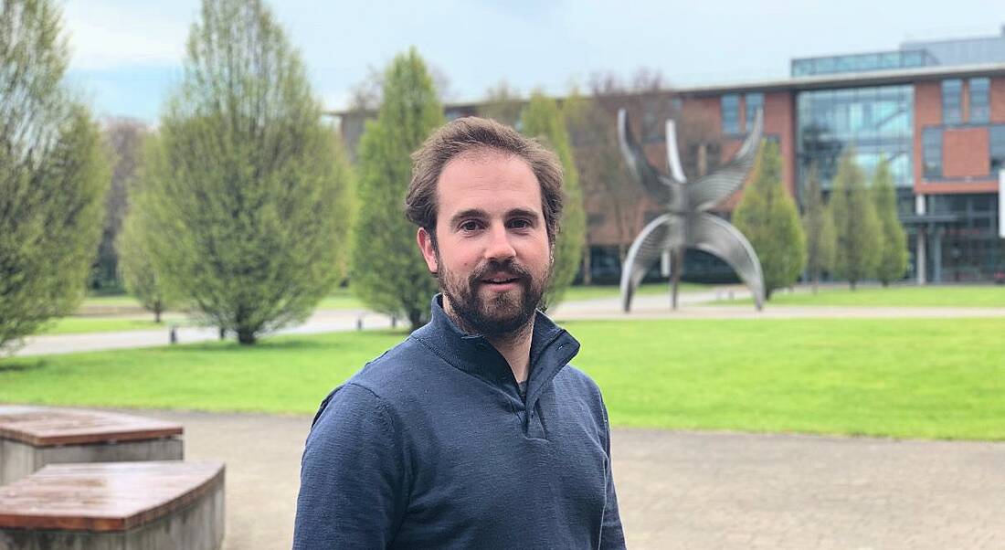 A dark-haired man with a beared wearing a navy jumper standing outside with grass behind him.