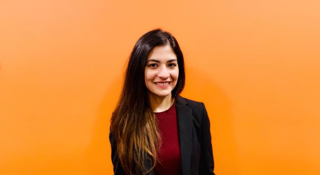 A young woman in a dark business suit standing against a vibrant orange wall smiling at the camera.