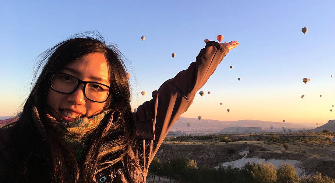 A close-up of a young woman with her arm outstretched behind her at hot air balloons. She works at New Relic.