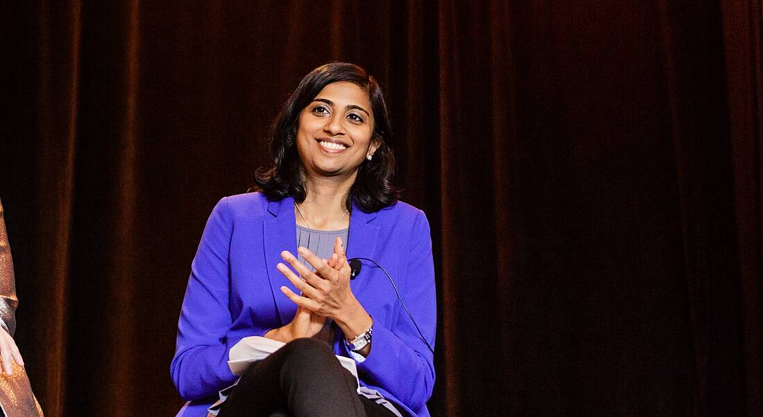 A young woman in a blue blazer sitting on a stage smiling. She’s speaking at a conference about the gender pay gap.