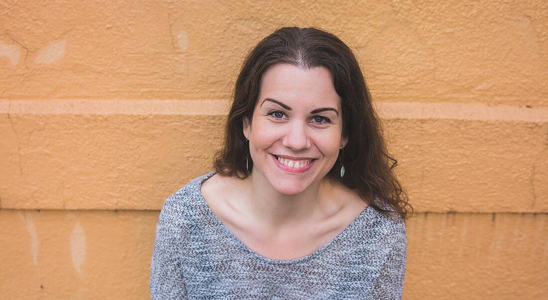 A woman with brown hair smiling against a bright orange wall. She is Jennifer Crow, an expert on grief in the workplace.