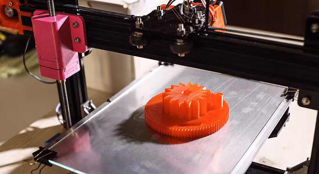 Image of an industrial 3D printer making a circular orange part during manufacturing process.