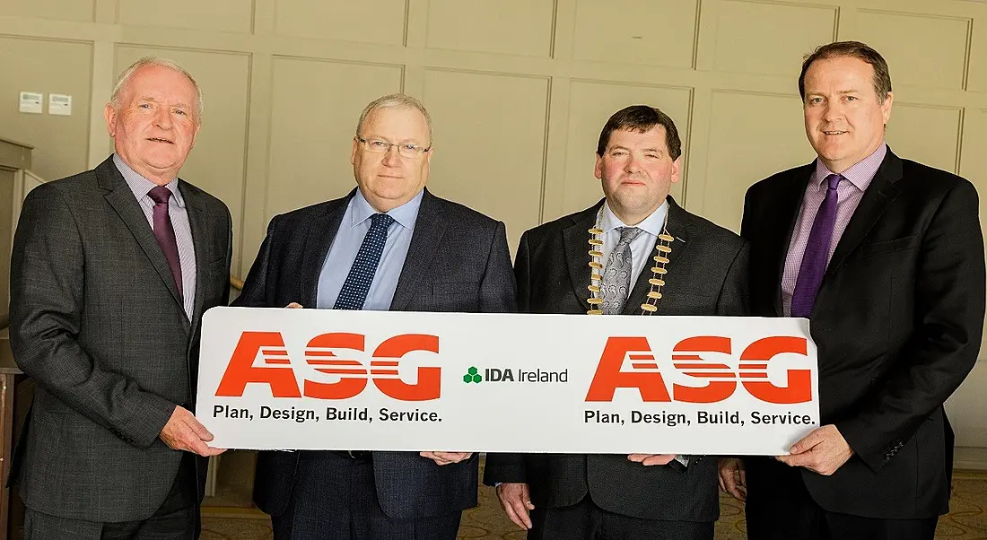 Four men in suits holding a long sign for AllState Sales Group against a beige wall.