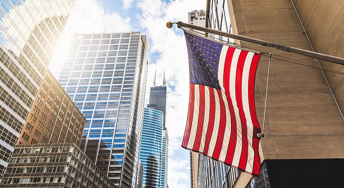 View from below an American flag billowing against the backdrop of a glittering, chrome metropolitan landscape.