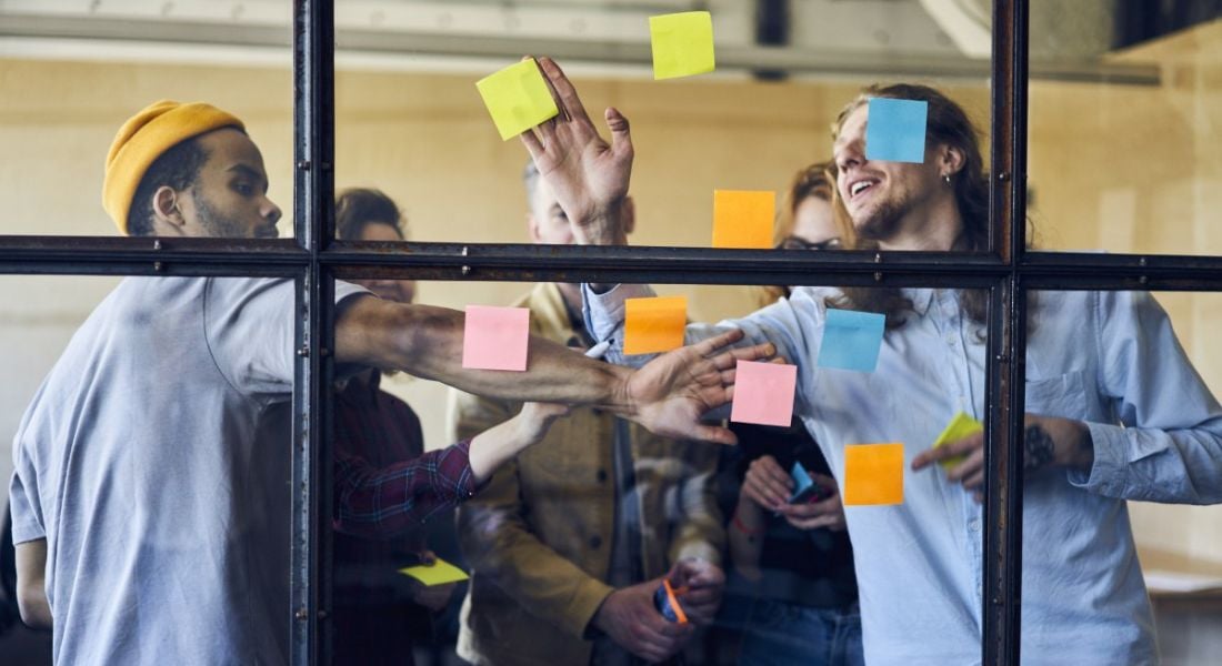 Group of young employees crowded around a window putting up coloured post-it notes and planning how to complete their latest task.
