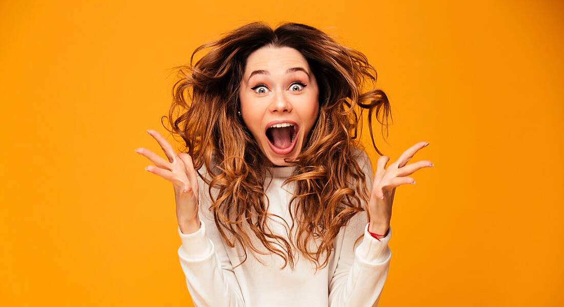 Woman jumping up with arms out in happy surprise with brunette curled tresses lifting into the air against orange background.