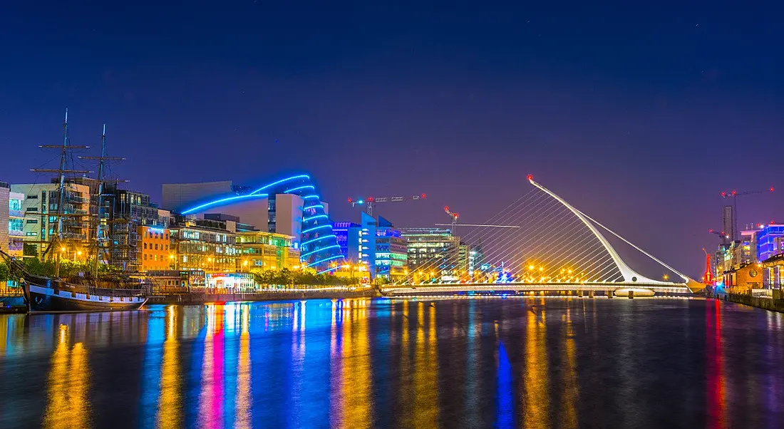 View of Dublin city’s Samuel Beckett bridge, Convention Centre and metropolitan skyline at night reflecting on the Liffey River.