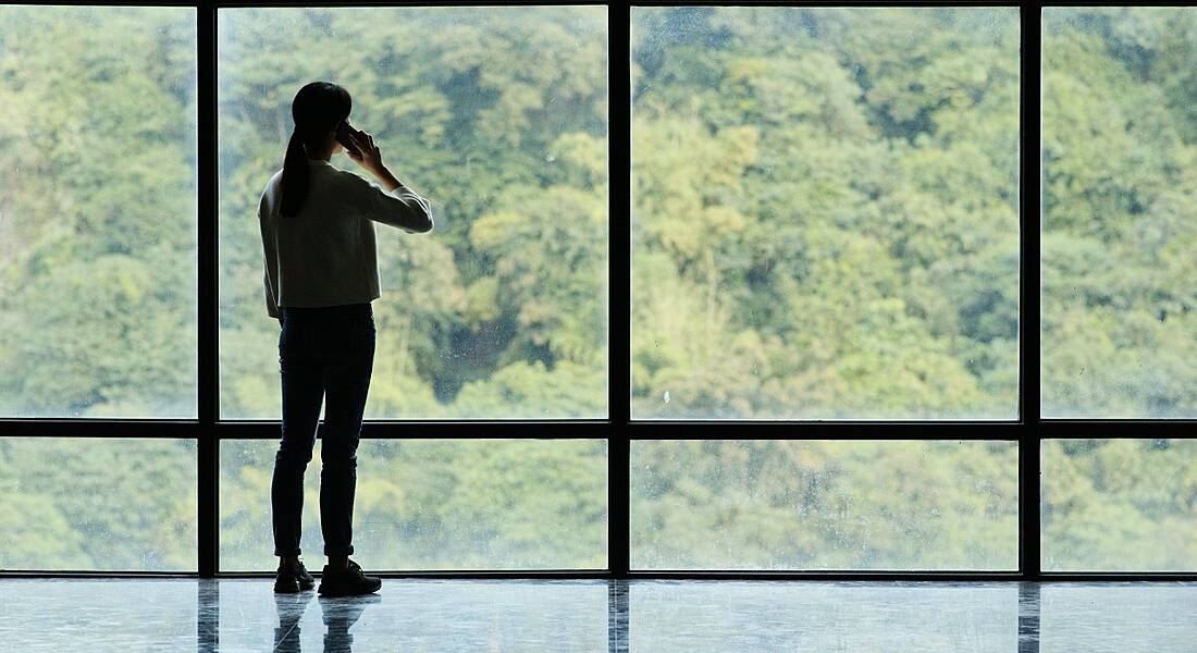 Silhouette of woman on phone looking at trees through tall ceiling-to-floor glass windows contemplating work-life balance.