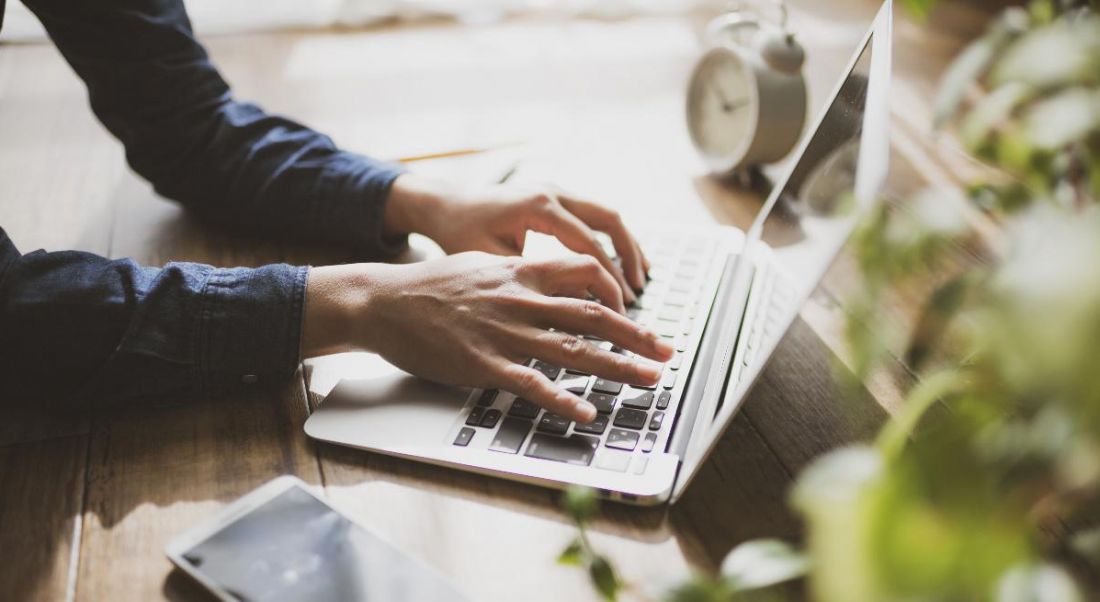 View of forearms clad in dark denim shirt and hands floating over the keyboard of a laptop.