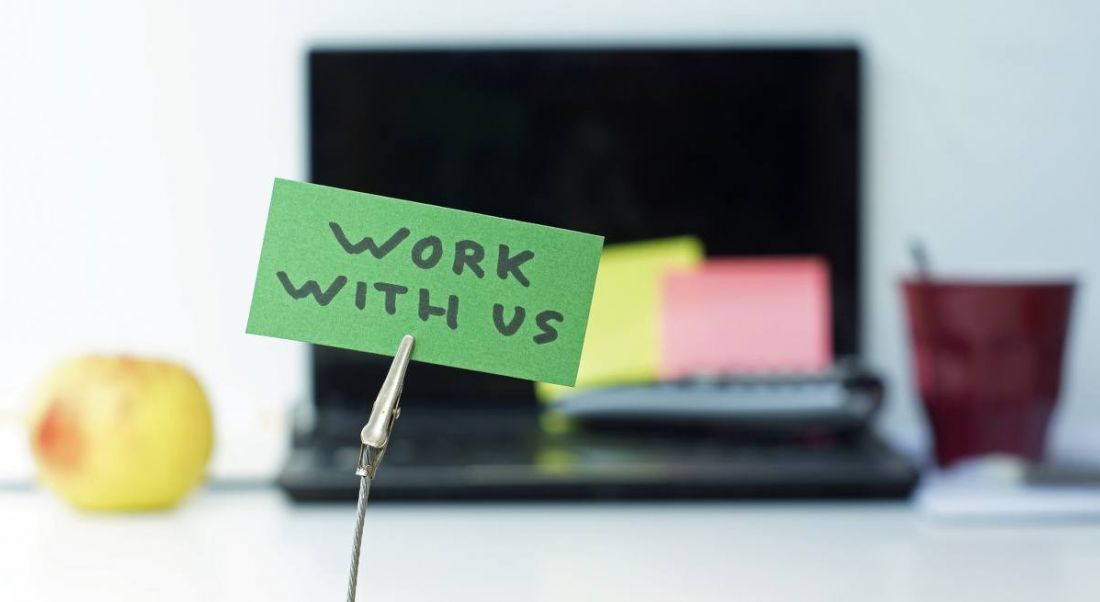 A clip holds a green note in place that reads ‘Work with us’. In the blurred background, we can see a laptop and coffee mug.