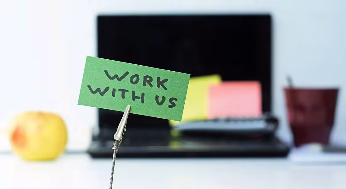 A clip holds a green note in place that reads ‘Work with us’. In the blurred background, we can see a laptop and coffee mug.