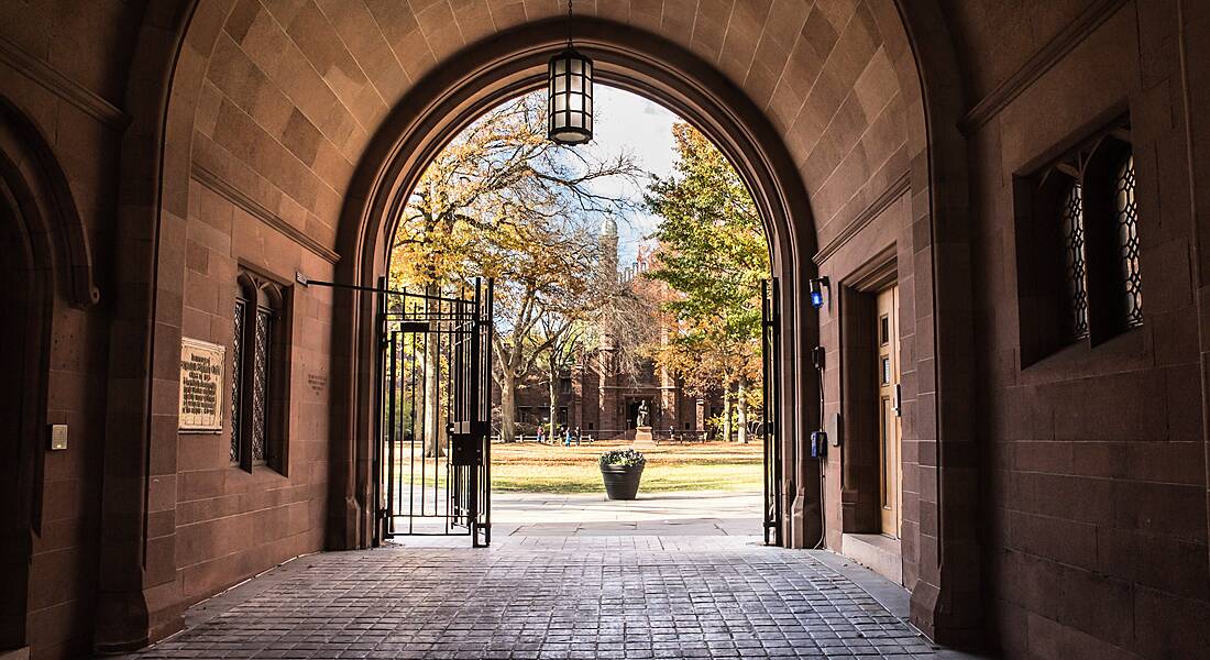 View of the gates at the end of an outdoor arched corridor looking out on to a green with distant people congregated on it.