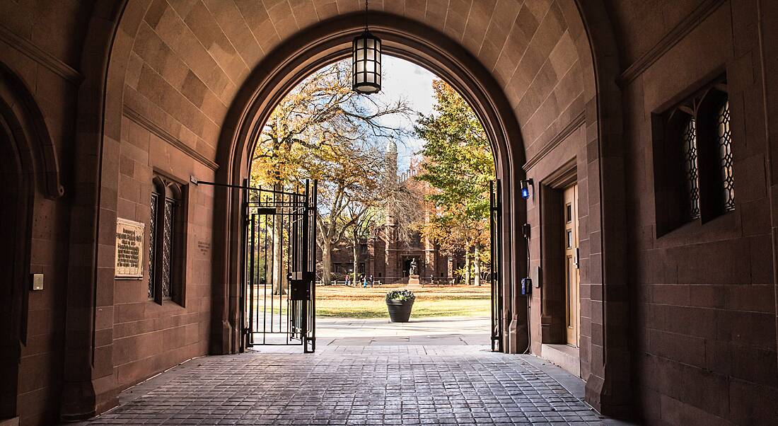 View of the gates at the end of an outdoor arched corridor looking out on to a green with distant people congregated on it.