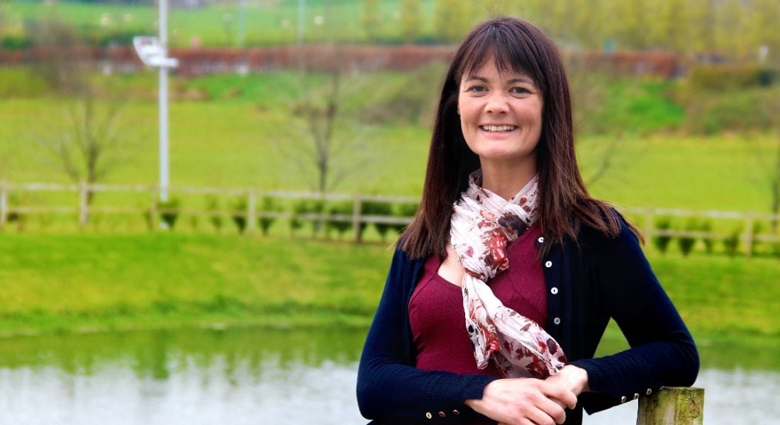 Brunette woman smiling and looking at the camera with her arms crossed against a bucolic green background.