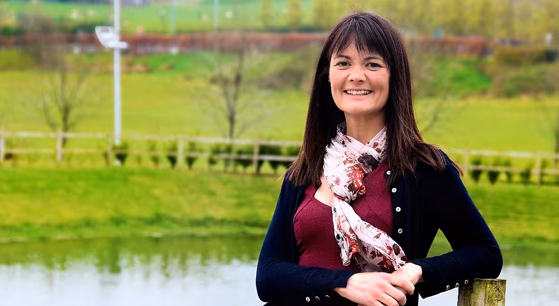 Brunette woman smiling and looking at the camera with her arms crossed against a bucolic green background.
