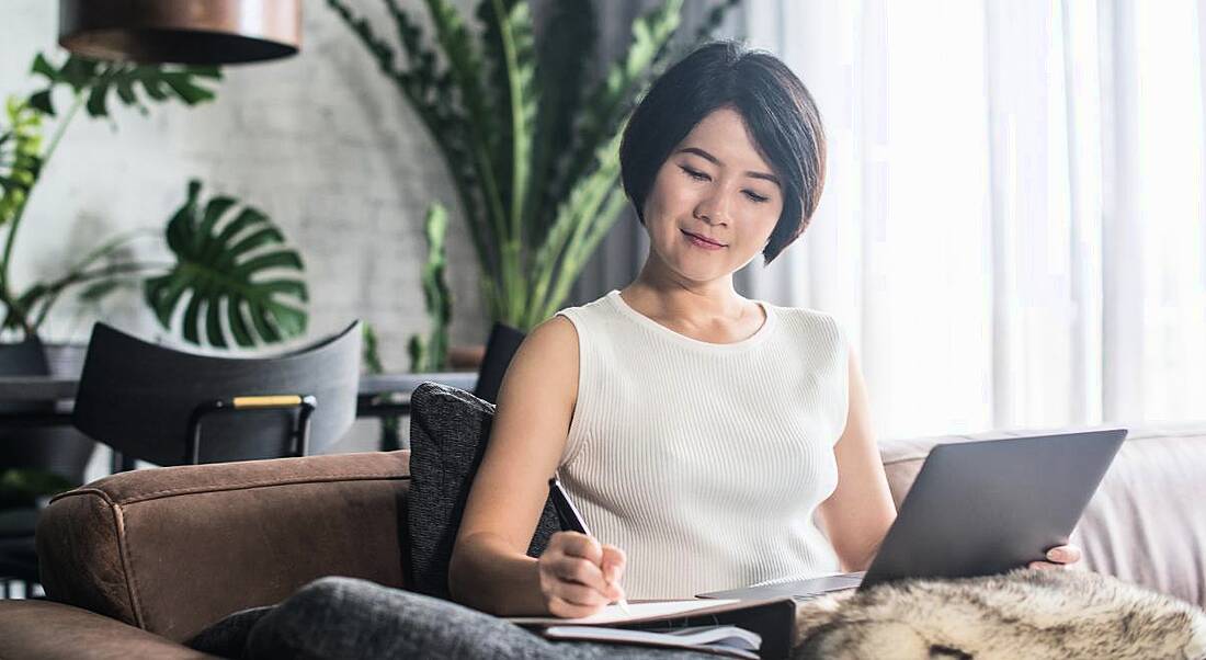 A woman sitting on a couch in her house making notes and working on a laptop, most likely updating her LinkedIn profile.