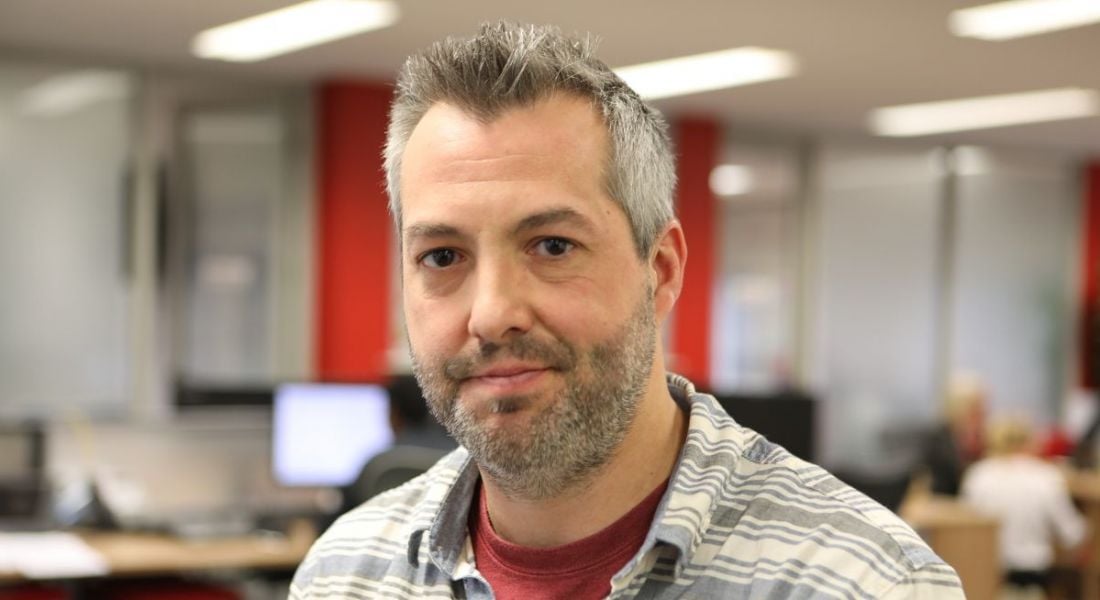 A view of a middle-aged men wearing a checked shirt looking serenely at camera against a red wall background.