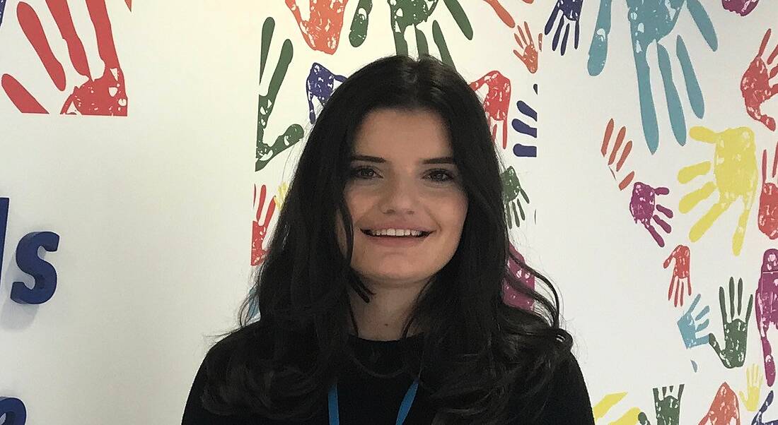 A young brunette woman smiling against a backdrop of a mural of multicoloured hand imprints.