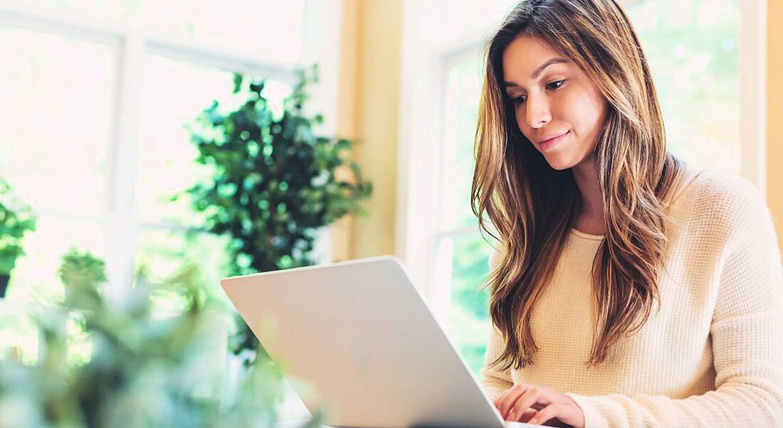 A woman in a warmly lit room sitting on a couch using a laptop. She is looking at the top fintech influencers.