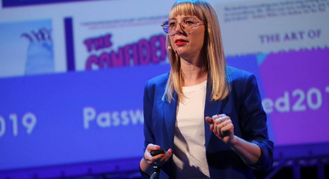Jennifer Romolini, a blonde woman with glasses wearing a blue blazer, on stage holding a screen remote control.