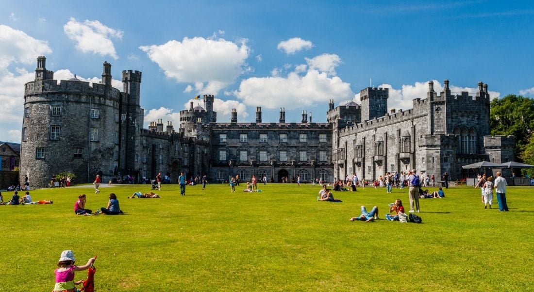 People sitting and playing on the grass in front of Kilkenny castle on a summer day under a blue sky.