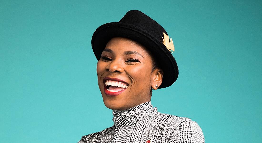 A close-up headshot of a smiling woman wearing a black hat against a bright turquoise background. She is Luvvie Ajayi.