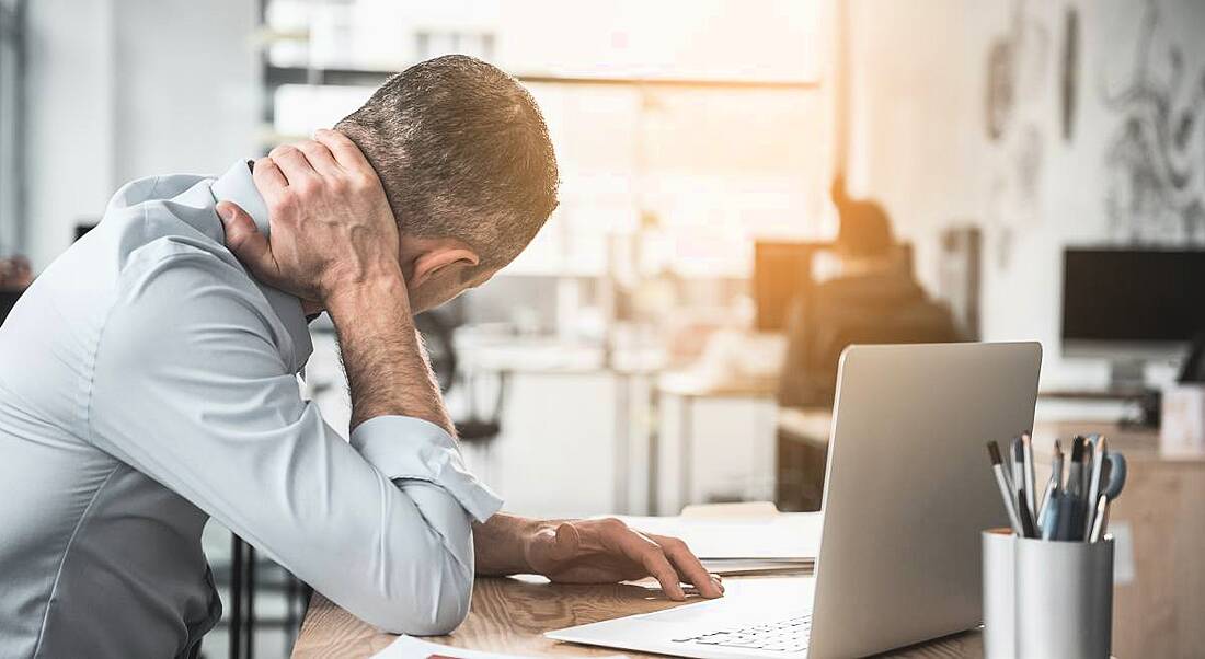 A man sitting at desk, holding the back of his neck, seemingly in pain, depicting someone at work with a long-term illness.