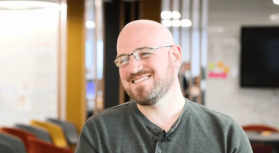 A bald man with a small beard wearing a causal green top and smiling. He works in software engineering.