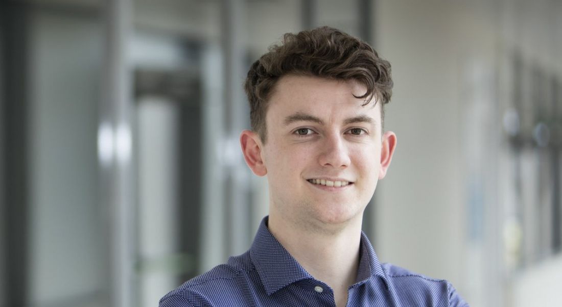 A close-up of a young man with dark hair wearing a dark blue shirt and smiling at the camera. He’s a graduate at Amgen.