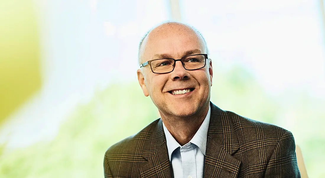 A bald, smiling man with glasses in a patterned brown suit against a bright background.