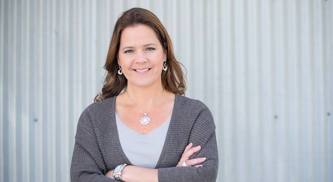 A brunette woman in a grey cardigan with arms crossed smiling at the camera. She is Susan Cooney.