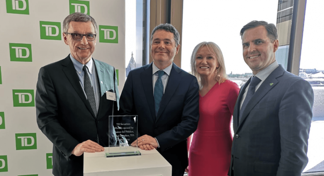 View of TD Securities green logo on background print with men and women in formal attire standing and smiling in front of it.