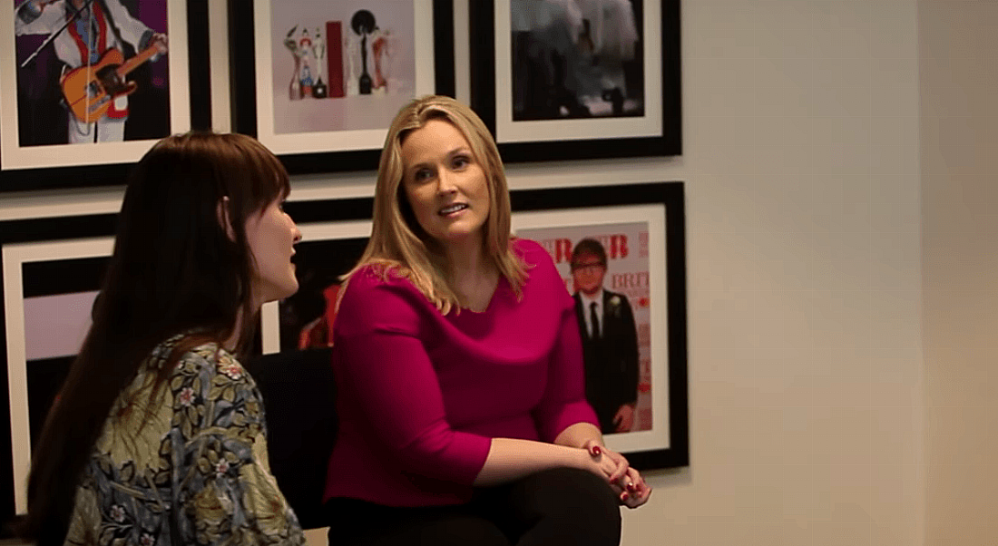 Two women sitting on low chairs turning in towards one another and talking against background of posters in dark frames.