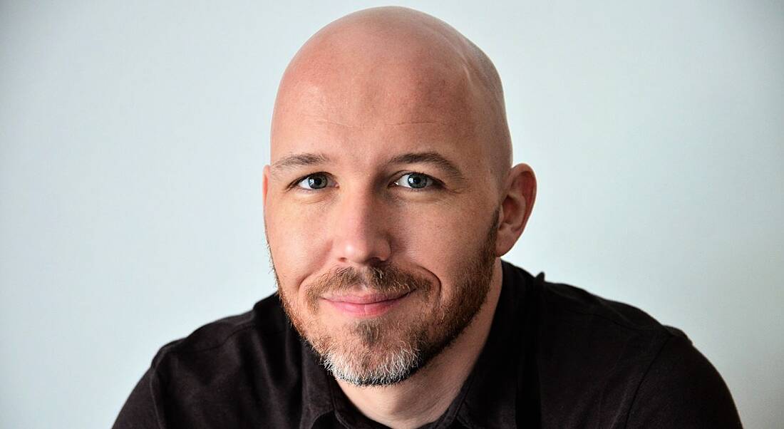 A close-up headshot of a bald man with stubble wearing a dark t-shirt. Here, he talks about paternity leave.