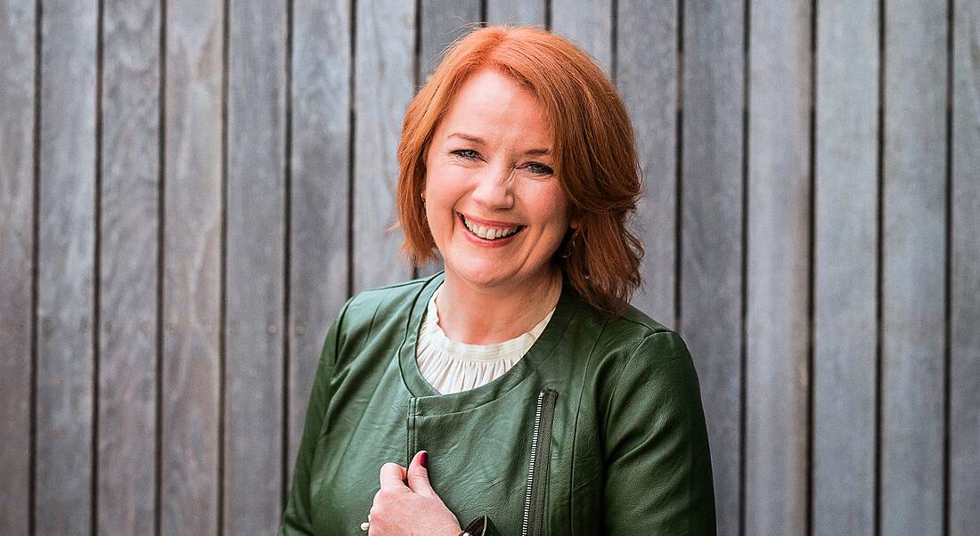 A woman with short red hair wearing a green jacket standing in front of a wooden fence.