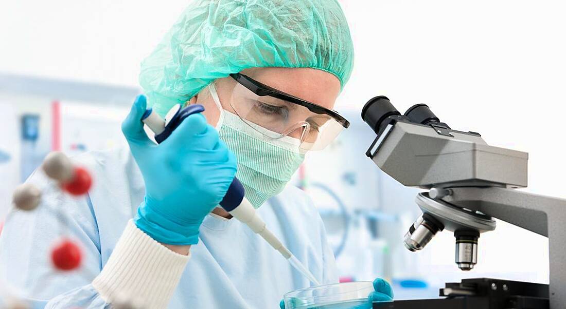 A scientist dressed in a lab coat, hair net and goggles, using a pipette near a microscope, depicting a life sciences career.