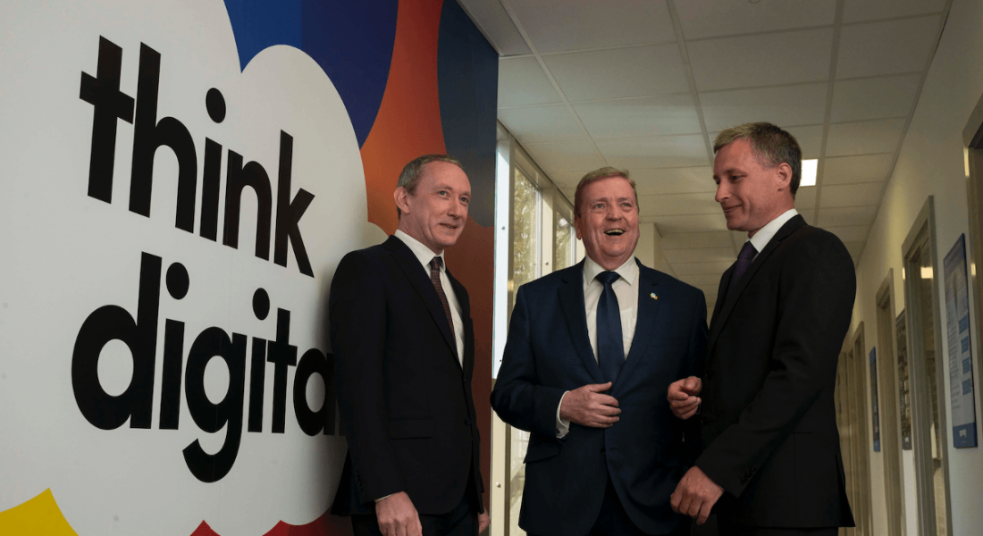 Three men in business suits standing and chatting and laughing next to a mural emblazoned with the words ‘think digital’.
