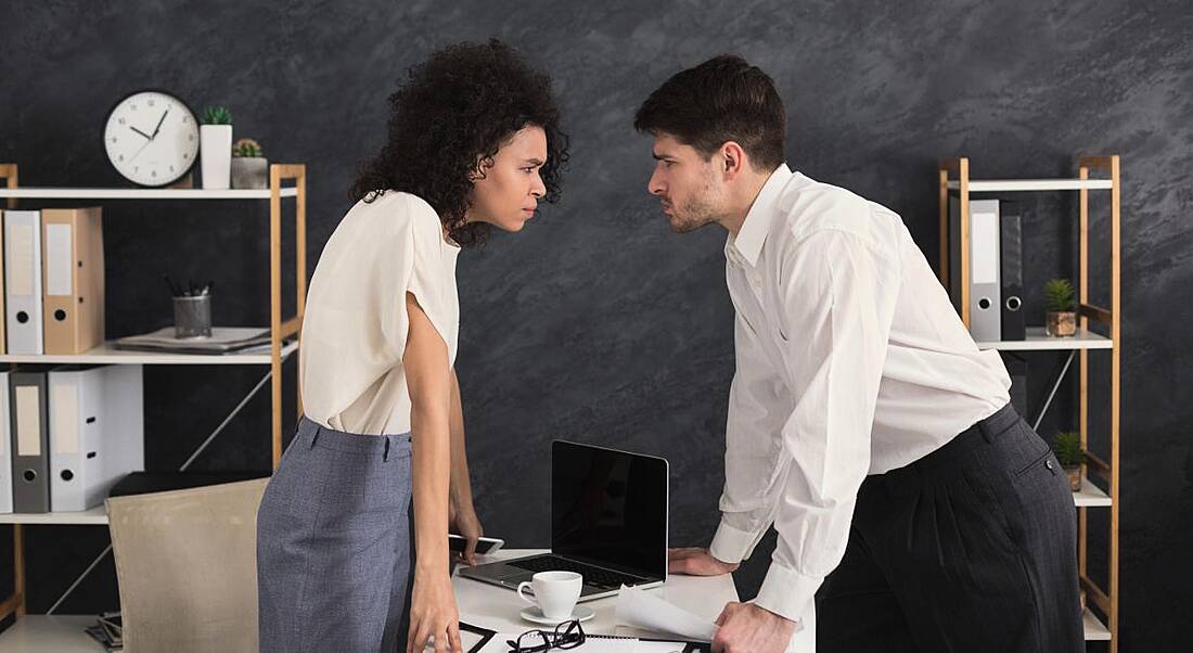 A male and female employee at logger heads lean into each other over an office desk with angry expressions.