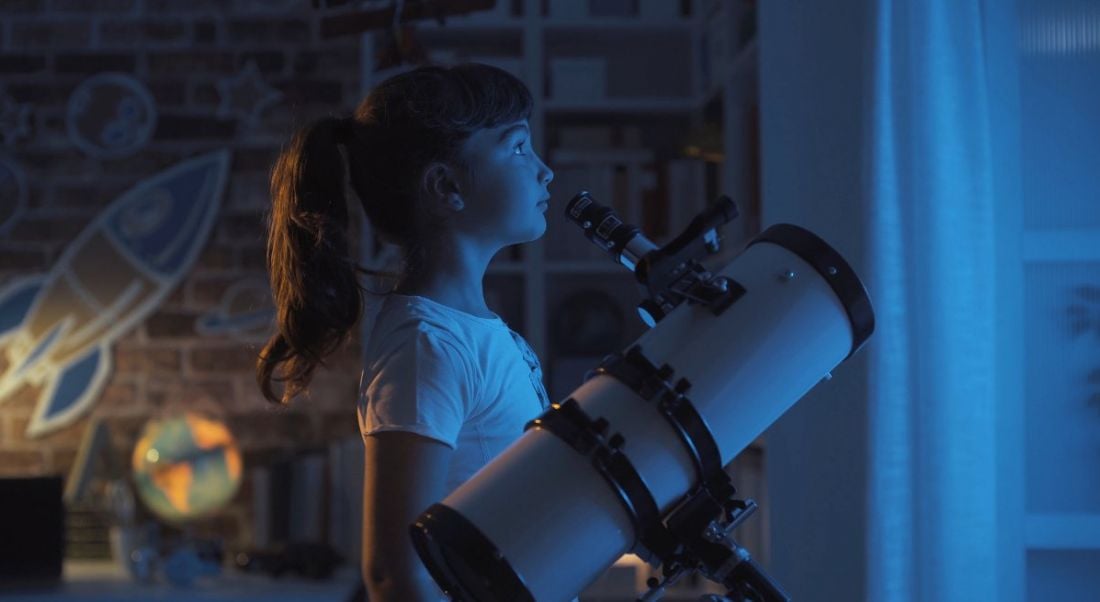 Young girl stargazing at night beside a telescope in a room with a globe and space decorations on the wall.