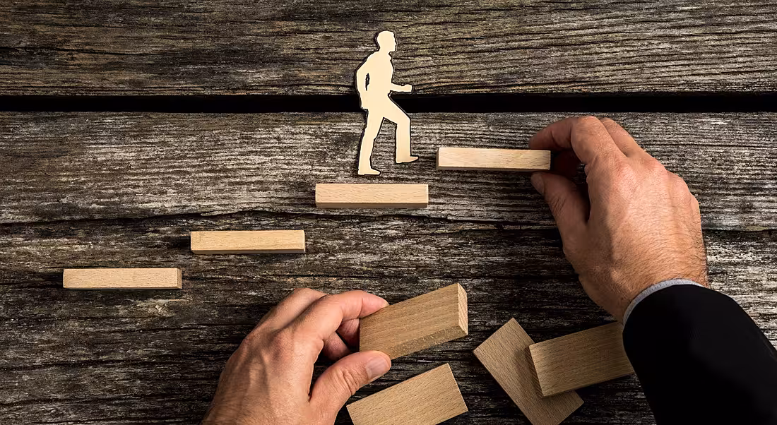 A wooden man flat on a table, walking on Jenga pieces being placed by human hands. It depicts someone getting promoted.
