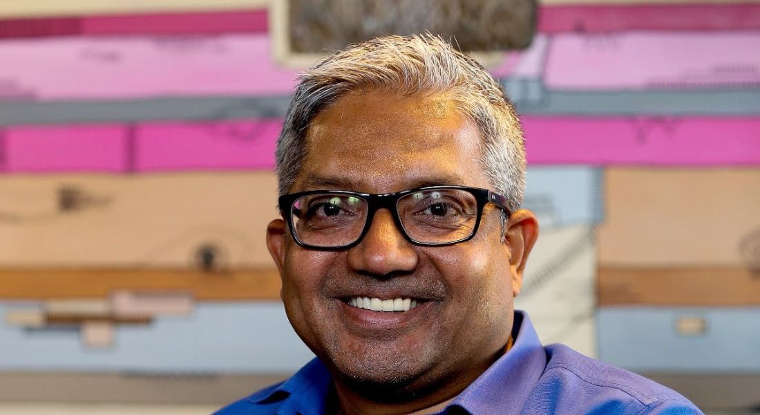 Smiling man with glasses wearing blue shirt and looking at camera, against striped backdrop.