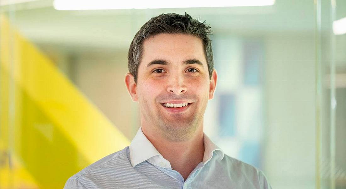 Man with salt and pepper hair looking straight at camera and smiling against a blurred multicoloured background.