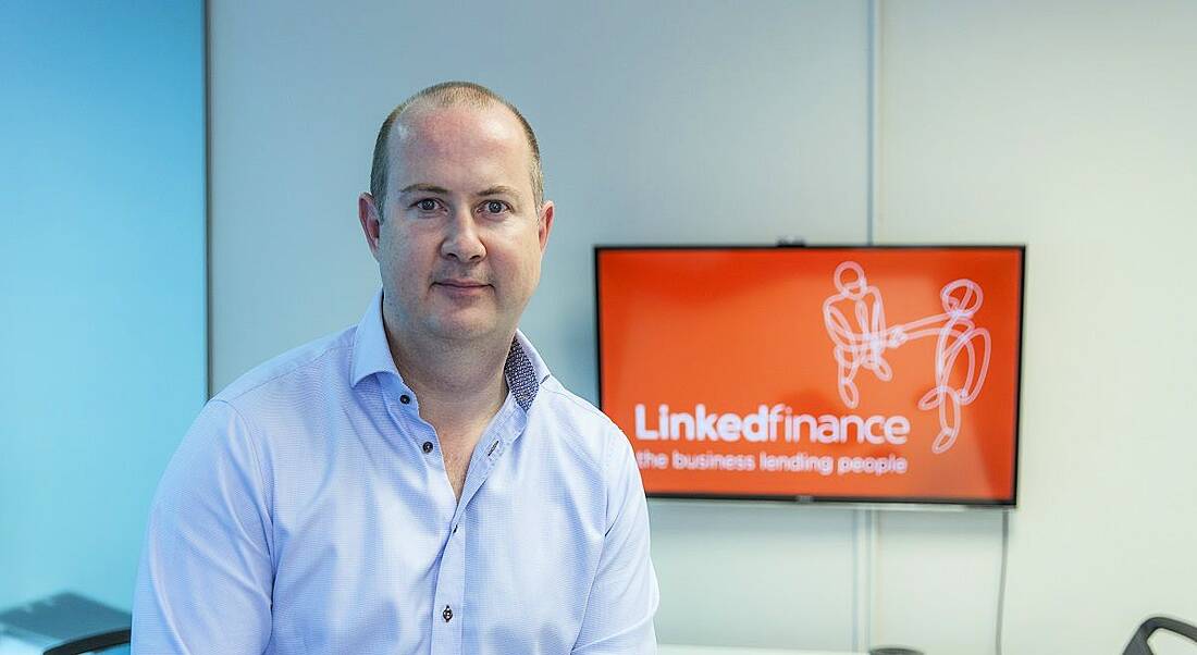 Niall Dorrian in a light blue shirt sitting on a desk with a Linked Finance logo behind him.