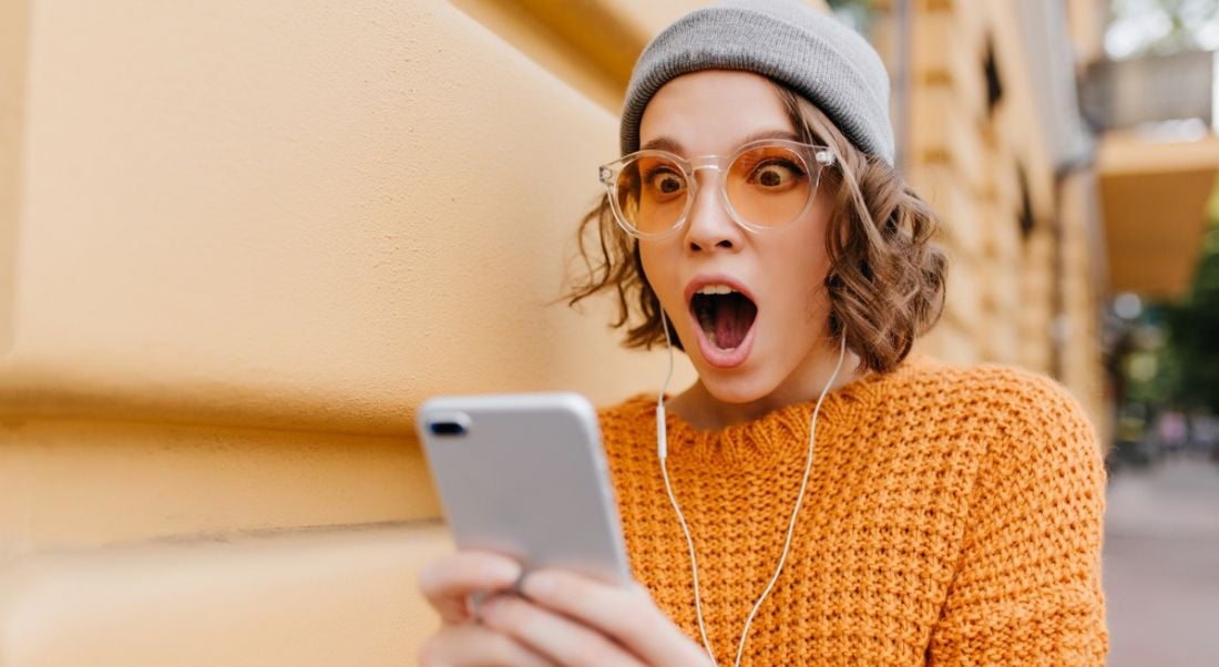 Woman with glasses, curly hair, grey beanie and orange sweater holding her phone and looking at it with awed expression.