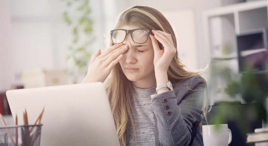 A young blonde woman lifting thick-rimmed glasses and rubbing her brow in consternation at her desk at work.