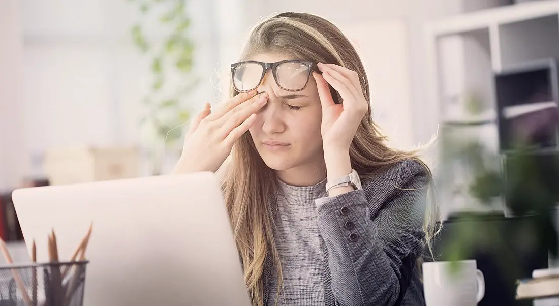 A young blonde woman lifting thick-rimmed glasses and rubbing her brow in consternation at her desk at work.