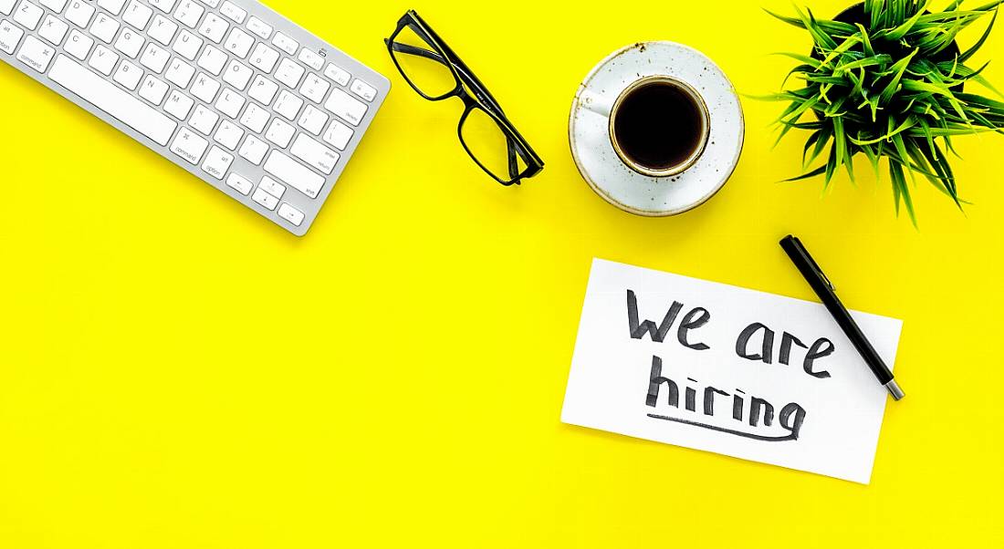 Looking down at a bright yellow desktop, where a ‘We are hiring’ sign sits next to a keyboard, a cup of coffee and a green plant.