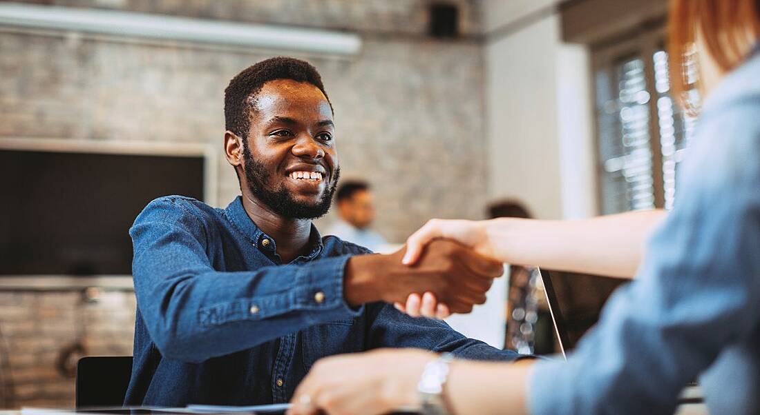 A young man in a denim shirt shakes the hand of a recruiter having successfully completed an interview.